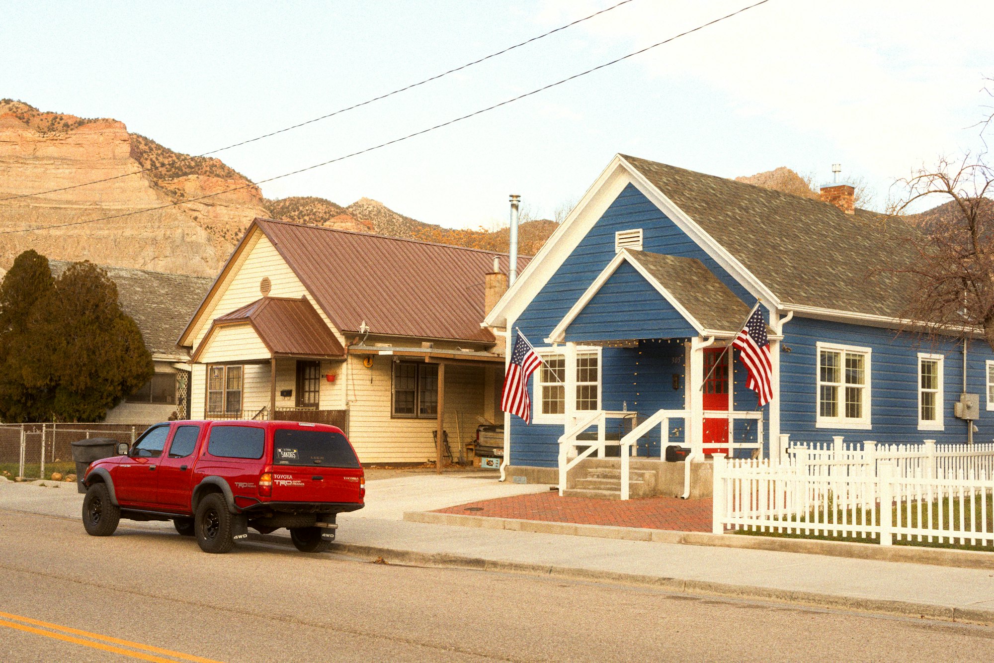 A blue house with a red door and a white picket fence, with red rock cliffs behind it.