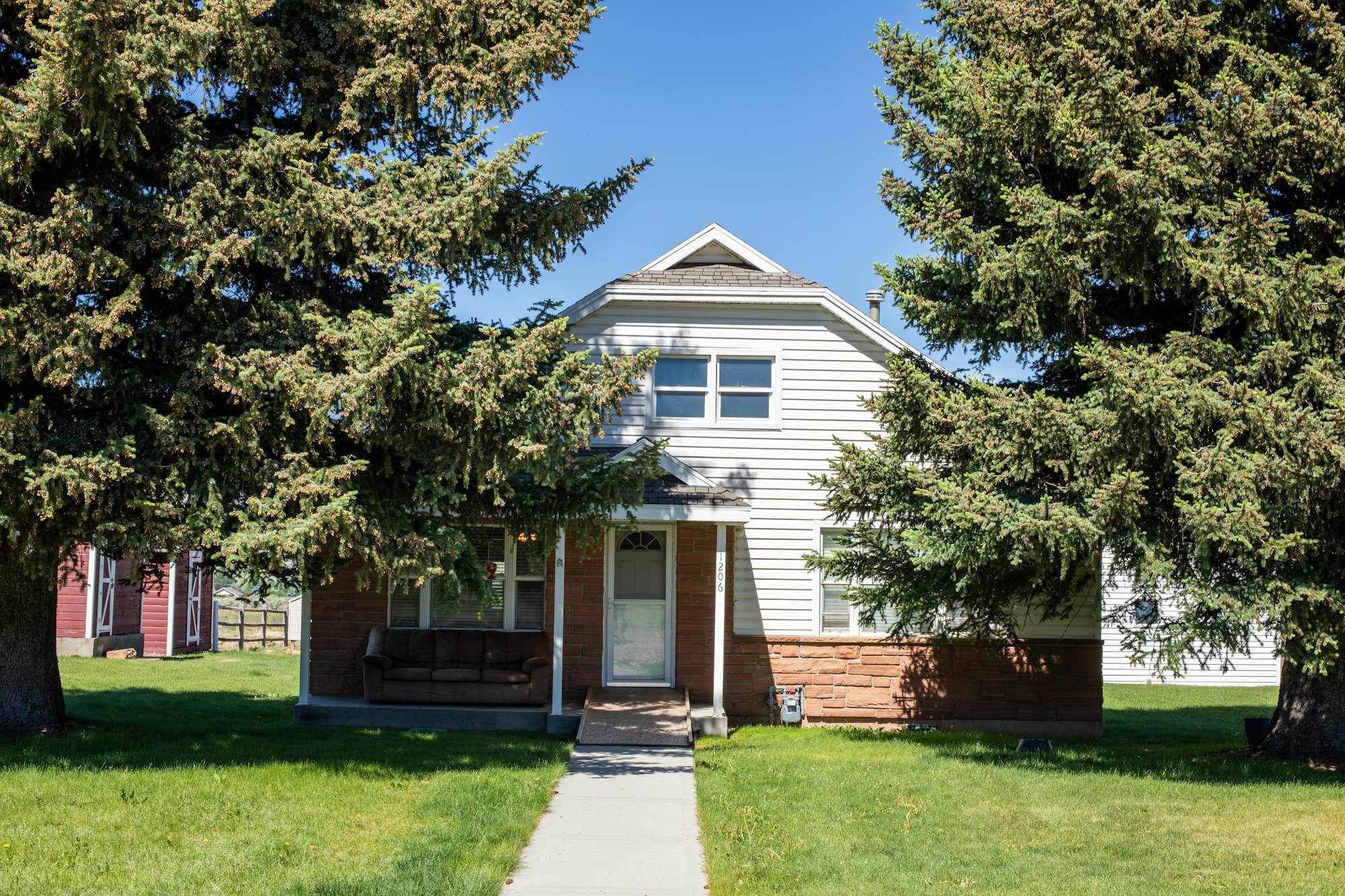 A white two-storey house with a brick base and a tall pine on a green lawn.